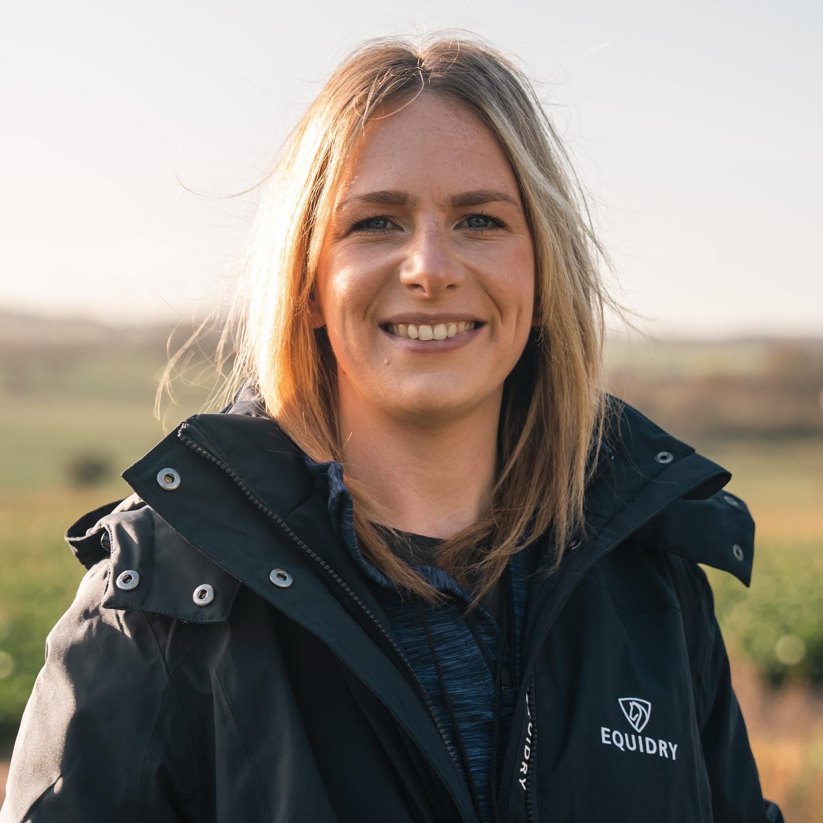 Katie crouching on a countryside path with two dogs in harnesses, smiling in golden hour light